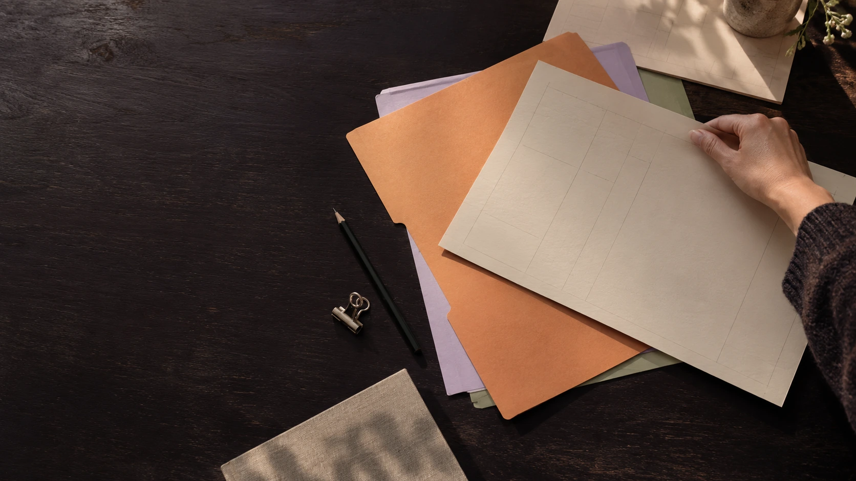 A hand arranging apricot, lilac and sage papers on a dark worktable.